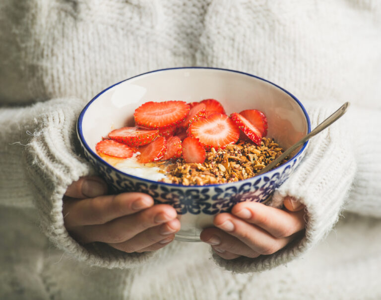 Healthy breakfast greek yogurt, granola and strawberry bowl in hands of woman wearing white loose woolen sweater, selective focus. Clean eating, healthy, vegetarian, dieting food concept