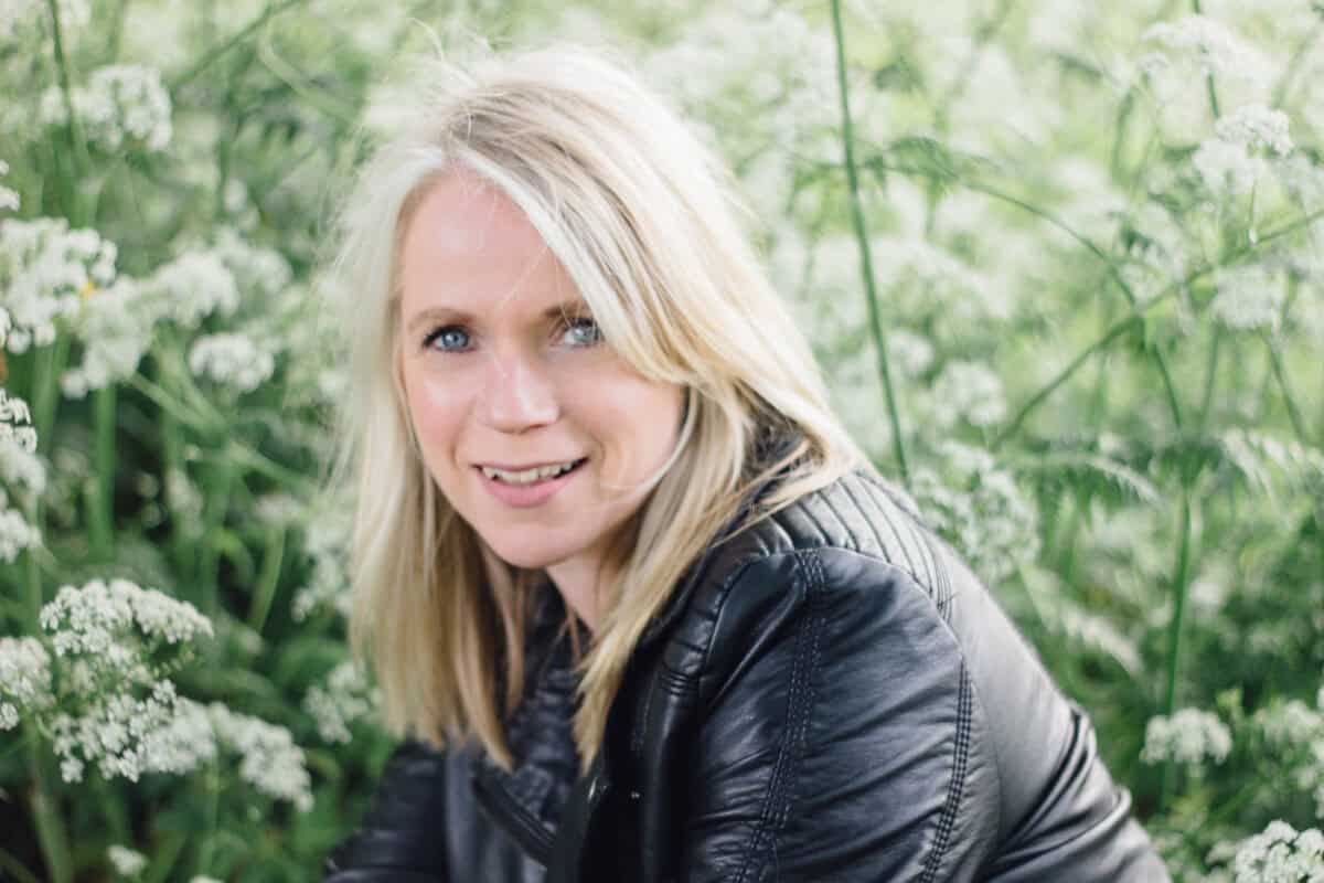 Een vrouw met blond haar en een zwart leren jack glimlacht terwijl ze buiten tussen hoge groene planten en witte wilde bloemen zit.