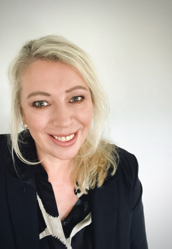 Een vrouw met lichtblond haar die een zwarte blazer en een blouse met patroon draagt, glimlacht naar de camera tegen een effen witte achtergrond.