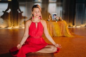 Een vrouw in een rode jurk zit in kleermakerszit op de grond te mediteren met gesloten ogen en ontspannen handen. Achter haar staat een klein altaar met ingelijste foto's en voorwerpen op een tafel die bedekt is met een oranje doek.