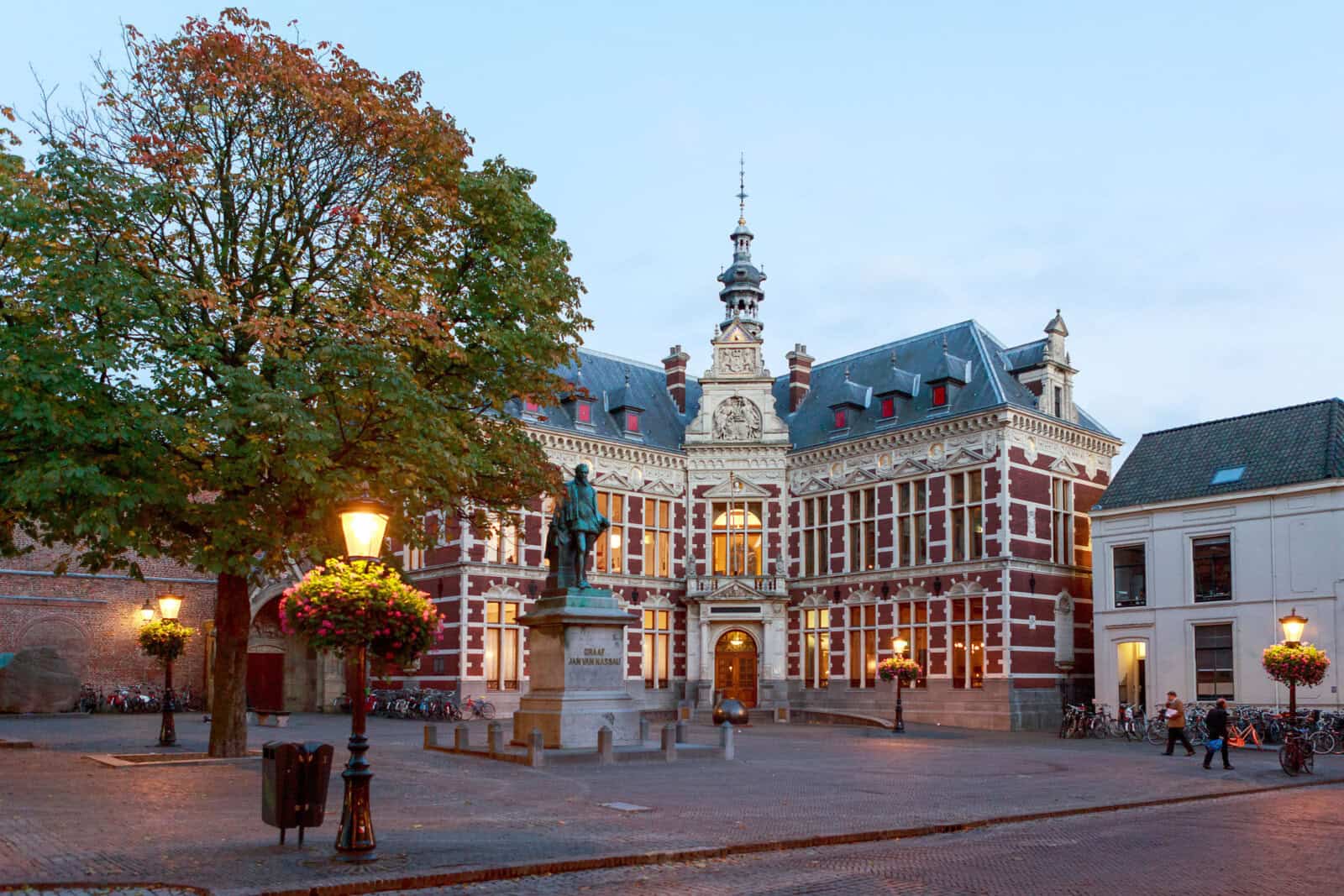 Historisch universiteitsgebouw met sierlijke architectuur en toren, gelegen op een met bomen omzoomd geplaveid plein in de schemering. Er staat een standbeeld voor, met straatverlichting en bloemenmanden die de omgeving verlichten.