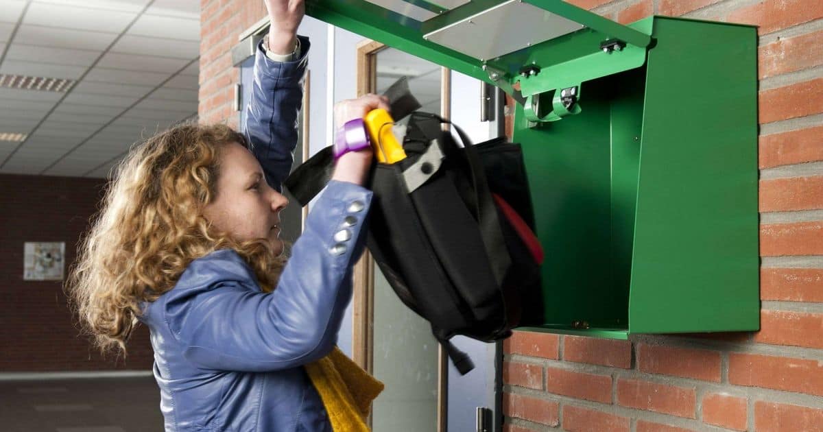 Een persoon met krullend haar en een blauwe jas plaatst zijn rugzak in een groen kastje dat aan een bakstenen muur in een gang is bevestigd.