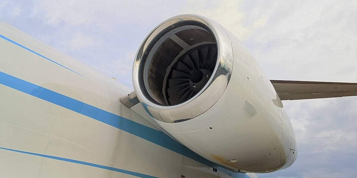 Een close-up van een straalmotor die aan de vleugel van een vliegtuig is bevestigd, waarbij een deel van de romp van het vliegtuig en blauwe strepen zichtbaar zijn tegen een bewolkte lucht.