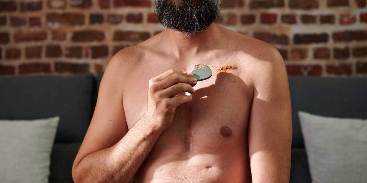 Een shirtloze man met baard gebruikt een elektrisch scheerapparaat om het haar van zijn borst te verwijderen terwijl hij op een bank voor een bakstenen muur zit.