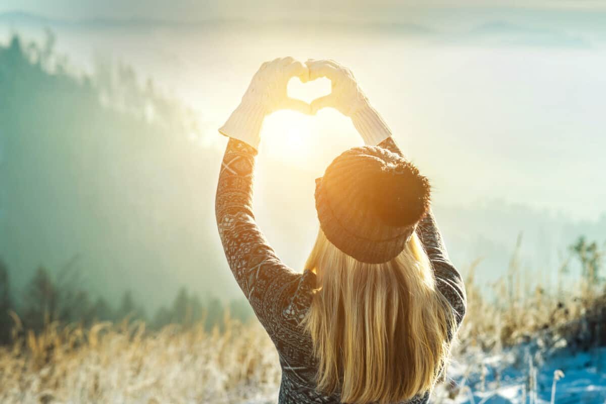 Happy traveler woman tourist stay on mountain top and making of love heart by hands in sunny frozen winter day.