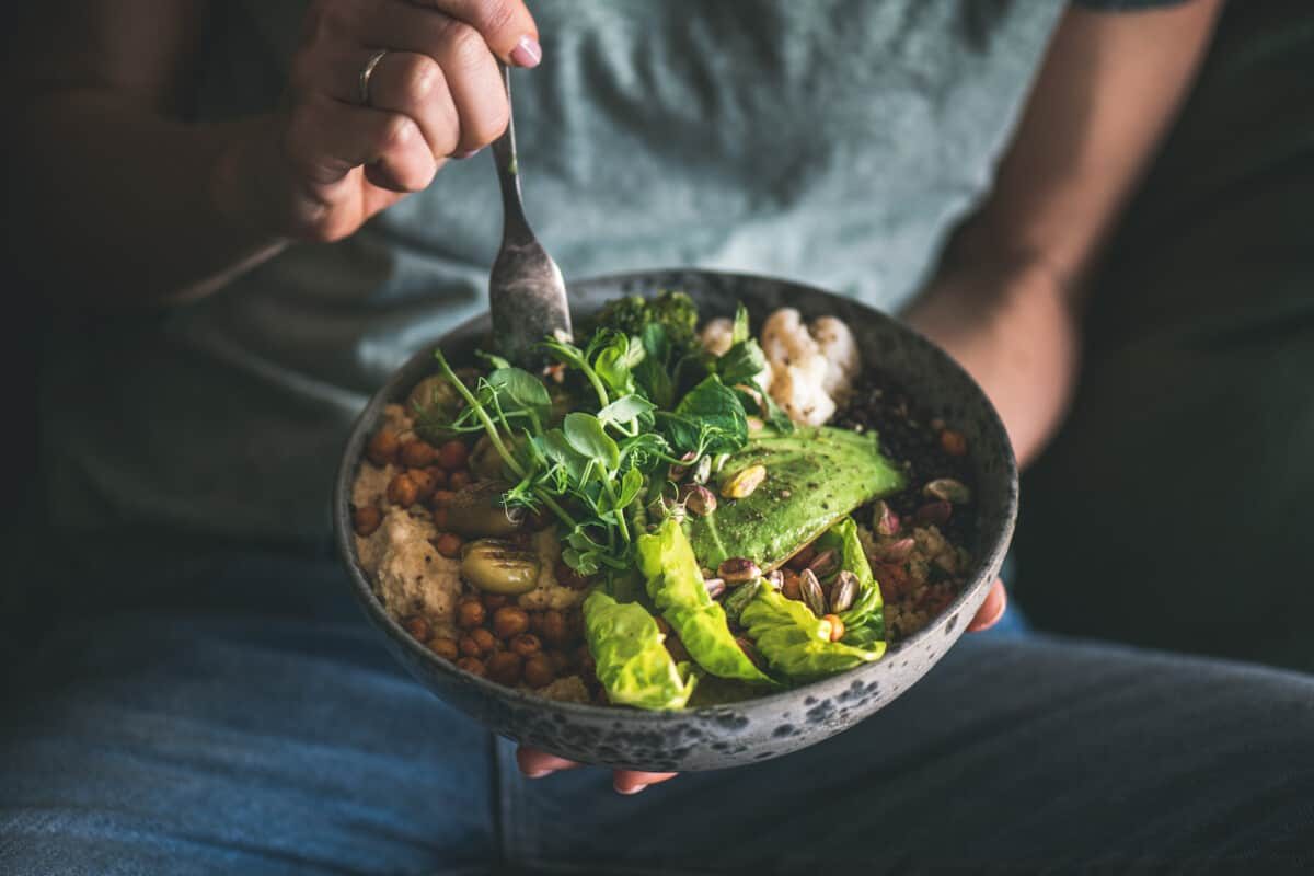 Healthy dinner or lunch. Woman in t-shirt and jeans eating vegan superbowl or Buddha bowl with hummus, vegetable, salad, beans, couscous and avocado