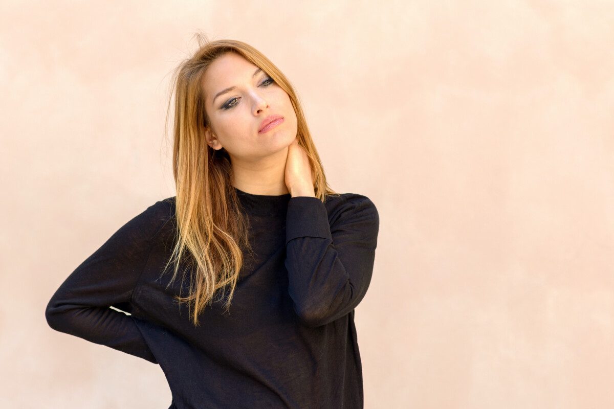 Natural portrait of a beautiful young woman with her hand on her neck and head tilted to the side gazing thoughtfully at the camera over a neutral studio background